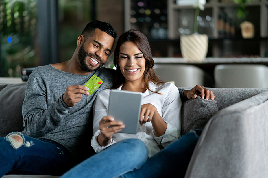 Happy couple sitting together on couch, man is holding debit card and woman is holding a tablet