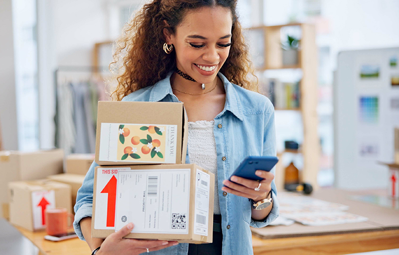 business woman smiling while holding cardboard packages and using a smartphone
